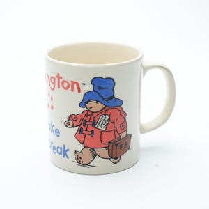 Op de afbeelding: Crèmekleurige keramische mok met een illustratie van Paddington Bear. De beer draagt een blauwe hoed en rode jas, met een bruine koffer. De mok heeft de woorden "Paddington" en "Take a break" erop.
