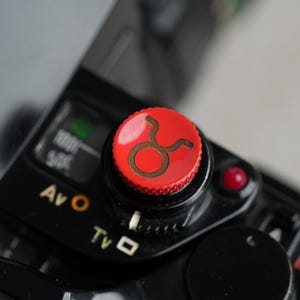 Peut inclure: Gros plan sur un appareil photo noir avec un bouton rouge sur lequel figure un symbole de taureau doré. L'appareil photo dispose de différents réglages, notamment "AV", "TV" et "O".