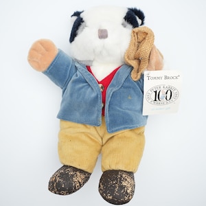 May include: A plush toy of Tommy Brock from Beatrix Potter's Peter Rabbit stories. The toy is wearing a blue jacket, red waistcoat, and yellow trousers. It is holding a brown bag in its right hand. The toy has a black and white face and a white nose. The tag on the toy reads "Tommy Brock" and "Peter Rabbit 100th Anniversary 1902-2002 Warne & Co."