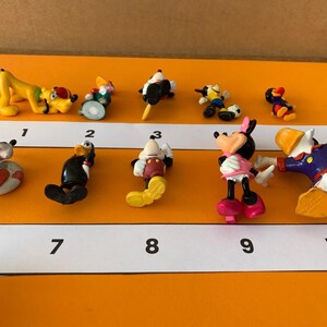 K&ouml;nnte beinhalten: Ein Set mit zehn kleinen Plastikfiguren von Disney-Charakteren, darunter Mickey Mouse, Minnie Mouse, Donald Duck, Goofy und Pluto. Die Figuren sind von 1 bis 10 nummeriert.
