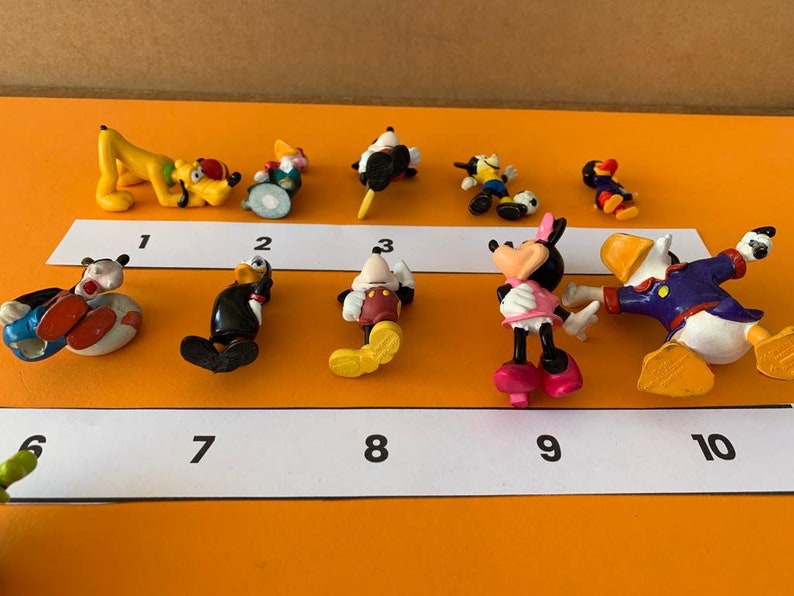 K&ouml;nnte beinhalten: Ein Set mit zehn kleinen Plastikfiguren von Disney-Charakteren, darunter Mickey Mouse, Minnie Mouse, Donald Duck, Goofy und Pluto. Die Figuren sind von 1 bis 10 nummeriert.