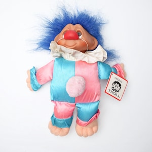 May include: A troll doll with bright blue hair, a red clown nose, and a white ruffled collar. The doll wears a satin-finish jumpsuit featuring a checkered pattern of light blue and pink panels, a pink pom-pom on the chest, and an attached tag reading ORIGINAL DAM TROLL.
