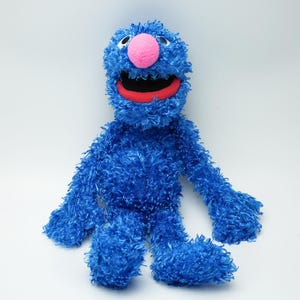 Könnte beinhalten: Ein blaues Plüschtier mit einer zotteligen, strukturierten Oberfläche. Die Figur hat eine große, runde, leuchtend rosa Nase, einen weit geöffneten Mund mit rotem Innenraum und zwei Augen mit schwarzen Pupillen. Arme und Beine sind lang und mit blauem Fell bedeckt.