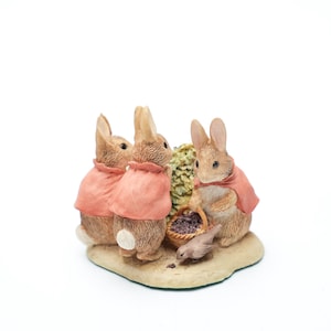 Op de afbeelding: Een beeldje met drie konijnen in roze capes, verzameld rond een mand met bessen. Eén konijn houdt de mand vast, een ander staat in de buurt. Een kleine vogel staat op de grond. De basis is beige.