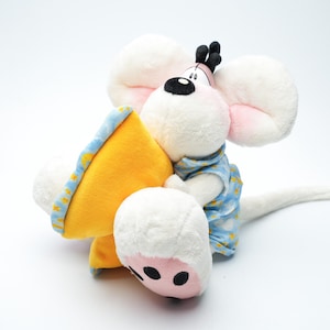 May include: A plush toy mouse with large white ears, a black nose, and small black antennae. The mouse is wearing a blue shirt and hugging a yellow wedge of cheese with blue trim. The toy is on a white background.