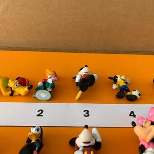 K&ouml;nnte beinhalten: Ein Set mit zehn kleinen Plastikfiguren von Disney-Charakteren, darunter Mickey Mouse, Minnie Mouse, Pluto und Donald Duck. Die Figuren sind von 1 bis 10 nummeriert und in einer Reihe auf einer wei&szlig;en Oberfl&auml;che angeordnet.