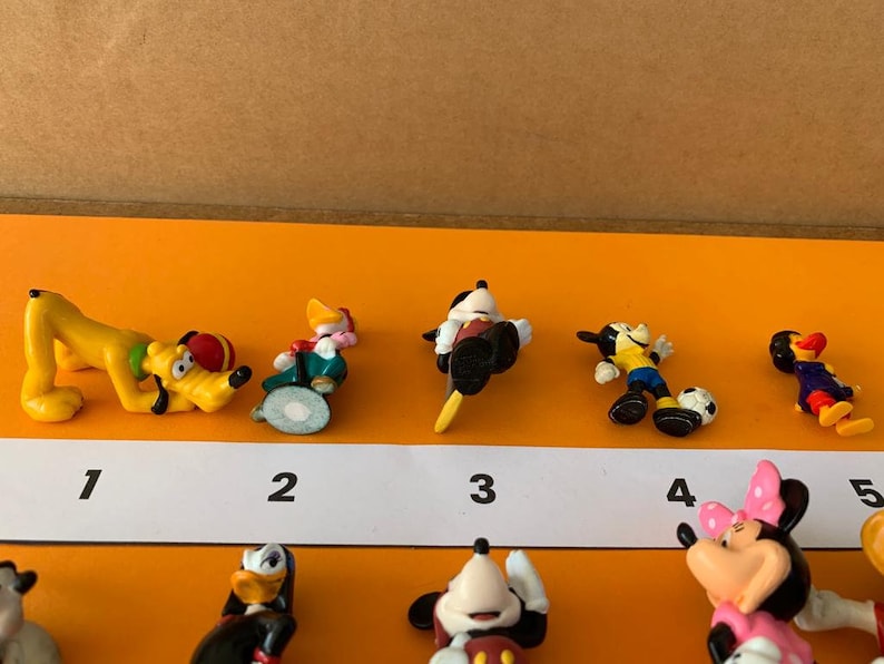 K&ouml;nnte beinhalten: Ein Set mit zehn kleinen Plastikfiguren von Disney-Charakteren, darunter Mickey Mouse, Minnie Mouse, Pluto und Donald Duck. Die Figuren sind von 1 bis 10 nummeriert und in einer Reihe auf einer wei&szlig;en Oberfl&auml;che angeordnet.