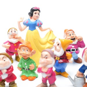 May include: A set of 11 plastic figurines featuring Snow White and the Seven Dwarfs. The figurines are brightly colored and feature detailed facial expressions. Snow White is wearing a yellow dress with a red bow and a blue bodice. The dwarfs are wearing various colored clothing and hats.