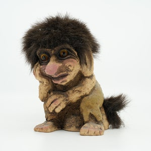 Könnte beinhalten: Eine braune und beigefarbene Trollfigur mit einem braunen, pelzigen Hut und Schwanz. Der Troll hat eine große Nase und große Augen.