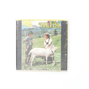 Könnte beinhalten: Eine Vintage-Filmspulenbox für 8mm-Film mit einer bunten Illustration von zwei Kindern mit einer Ziege auf einer Wiese. Die Box ist mit "Heidi" und "Une Exclusivité Avo Film Milano-Italy" beschriftet.