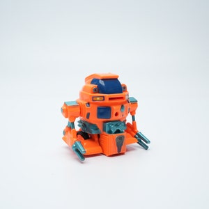 Könnte beinhalten: Orangefarbener und blauer Spielzeugroboter mit beweglichen Armen und Beinen. Der Roboter hat einen runden Körper und ein blaues Visier.