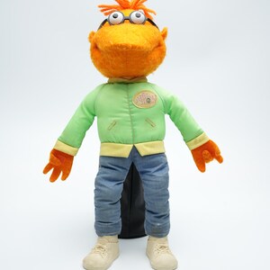 Peut inclure: Marionnette en peluche orange avec des cheveux roux, portant une veste verte avec un patch jaune qui dit "The Muppets", un jean bleu et des baskets blanches.