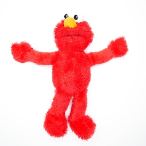Może przedstawiać: Czerwona pluszowa zabawka Elmo z ulicy Sezamkowej z żółtym nosem i dużymi białymi oczami. Zabawka leży na plecach z wyciągniętymi ramionami.
