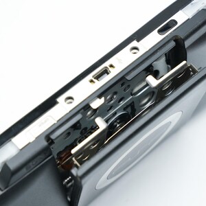 May include: Close-up of a black handheld gaming console with a silver trim. The console's open compartment reveals internal components and a USB port. The device is set against a white background.