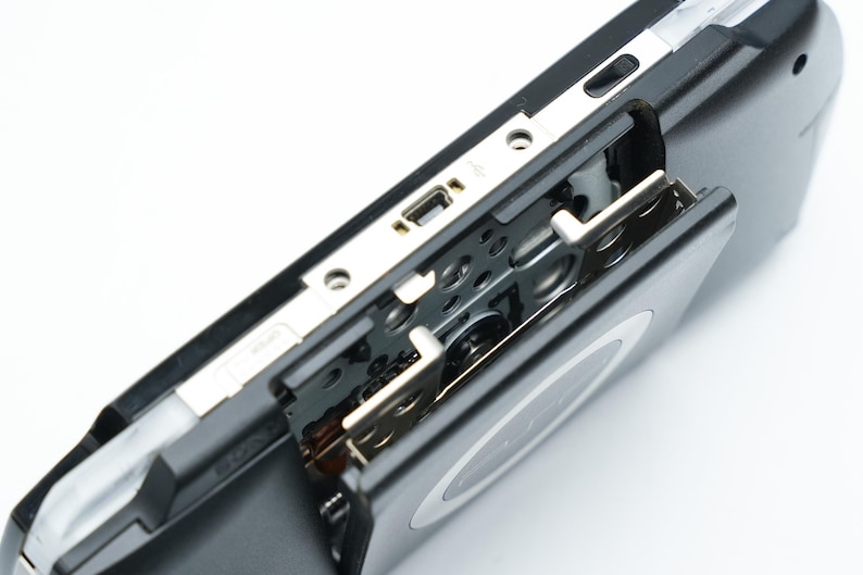 May include: Close-up of a black handheld gaming console with a silver trim. The console's open compartment reveals internal components and a USB port. The device is set against a white background.