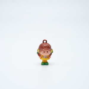 May include: A small plastic figurine of a girl with brown hair, wearing a yellow dress and green shoes. She has a pink face and yellow flowers in her hair.