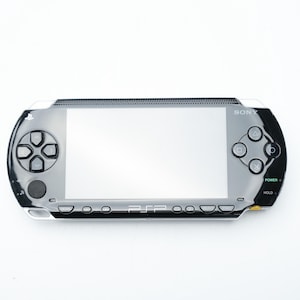 Puede incluir: Una consola de juegos portátil Sony PSP plateada y negra. El dispositivo cuenta con una pantalla grande, un panel direccional, botones de acción y un botón de encendido. La PSP está diseñada para juegos y entretenimiento portátiles.