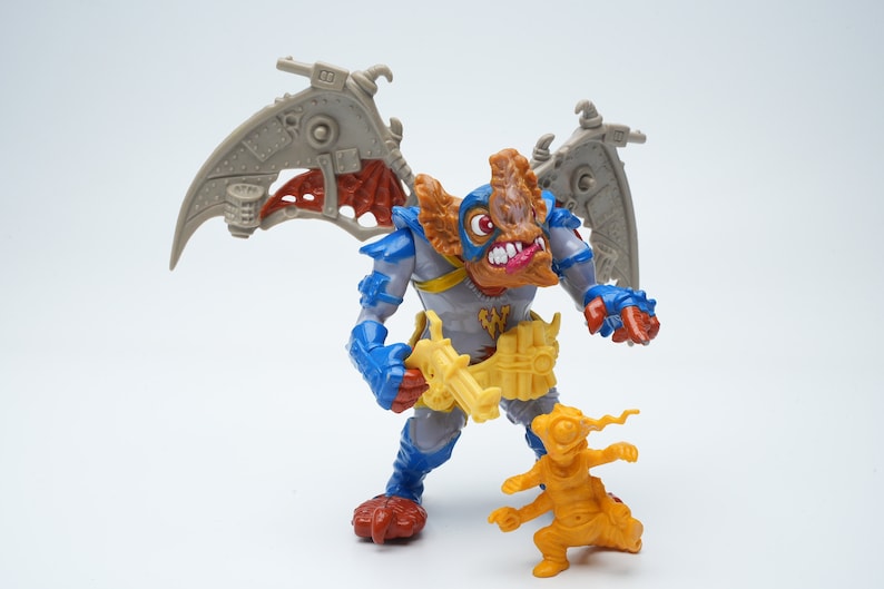 May include: A gray, blue, and orange action figure with wings and a gun. The figure is standing next to a smaller, yellow action figure.