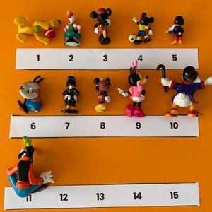 K&ouml;nnte beinhalten: Eine Sammlung von 15 kleinen Plastikfiguren verschiedener Disney-Charaktere, darunter Mickey Mouse, Minnie Mouse, Donald Duck, Goofy und Pluto. Die Figuren sind in Reihen auf einer wei&szlig;en Oberfl&auml;che angeordnet, wobei sich unter jeder Figur die Zahlen 1 bis 15 befinden.
