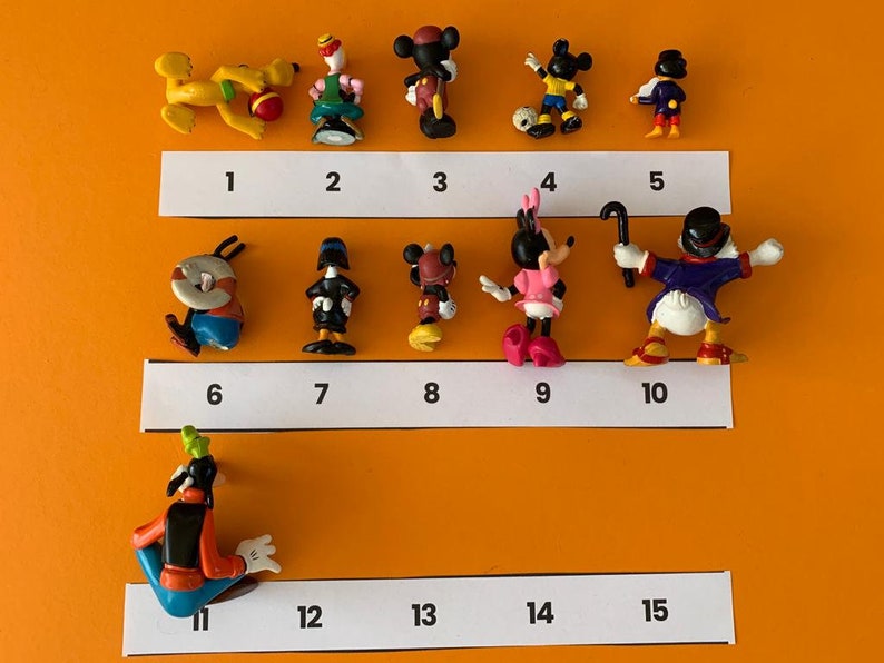 K&ouml;nnte beinhalten: Eine Sammlung von 15 kleinen Plastikfiguren verschiedener Disney-Charaktere, darunter Mickey Mouse, Minnie Mouse, Donald Duck, Goofy und Pluto. Die Figuren sind in Reihen auf einer wei&szlig;en Oberfl&auml;che angeordnet, wobei sich unter jeder Figur die Zahlen 1 bis 15 befinden.