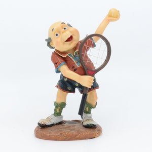 May include: A figurine of a man playing tennis. He is wearing a red and blue shirt, blue shorts, and green socks. He is holding a tennis racket and a yellow tennis ball. The figurine is standing on a brown base.