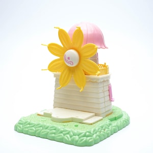 May include: A yellow flower windmill toy with a pink roof and a green base. The toy is made of plastic and has a whimsical design.