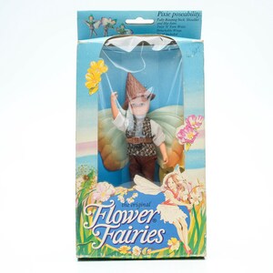 Op de afbeelding: Een Flower Fairies pop in een doos met een bruine hoed en vleugels. De pop houdt een gele bloem vast. De doos is blauw en wit met de tekst "The Original Flower Fairies" erop gedrukt.