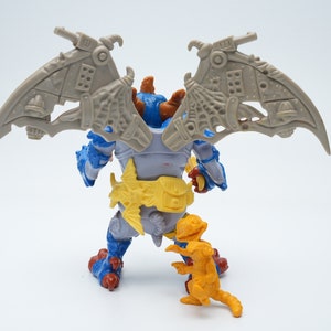 May include: A blue and gray action figure with wings and a yellow belt. The figure is standing on a white surface with a smaller orange figure in front of it.