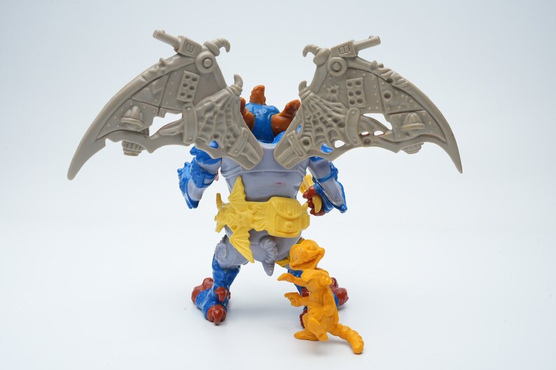 May include: A blue and gray action figure with wings and a yellow belt. The figure is standing on a white surface with a smaller orange figure in front of it.