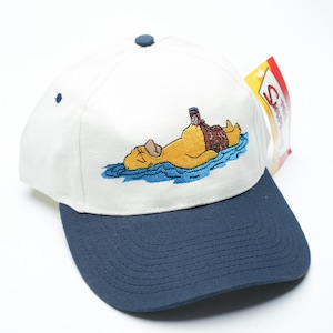 Op de afbeelding: Een witte en marineblauwe baseballcap met een geborduurd beeld van een gele figuur die in blauw water met een biertje drijft. De cap heeft een gebogen klep en een kleine knoop bovenop. Een label is aan de zijkant bevestigd.