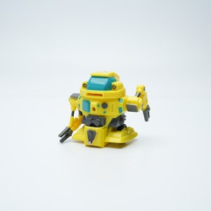 Könnte beinhalten: Ein gelbes und türkisfarbenes Spielzeugroboter mit schwarzen Armen und Beinen. Der Roboter hat einen runden Körper mit einem flachen Boden und einen kleinen, runden Kopf.