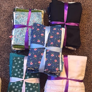 May include: Five stacks of folded fabric tied with satin ribbons. The stacks include a teal paisley print, a solid black fabric, a dark blue floral print, a dark green fabric with white snowflakes, and a white fabric with horizontal lace trim.