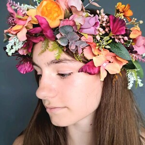 May include: A floral crown with pink, orange, and green flowers. The crown is made of silk flowers and is worn by a person with long brown hair.