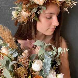 May include: A young person wearing a flower crown made of dried flowers and greenery. The crown is brown, beige, and green. The person is holding a bouquet of white, beige, and orange flowers with greenery.