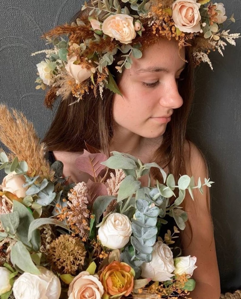 May include: A young person wearing a flower crown made of dried flowers and greenery. The crown is brown, beige, and green. The person is holding a bouquet of white, beige, and orange flowers with greenery.