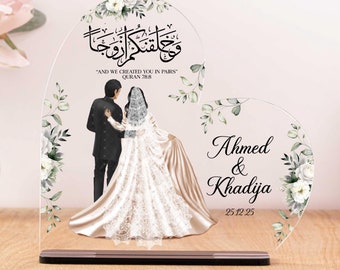May include: A heart-shaped acrylic wedding keepsake with a white floral design. The couple is depicted in black and white, with the bride wearing a white dress and the groom wearing a black suit. The Arabic text reads "And We Created You in Pairs" (Quran 78:8). The names Ahmed & Khadija and the date 25.12.25 are also included.