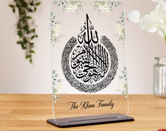 Può includere: Una placca in acrilico trasparente con un designo di calligrafia nera della frase islamica "Allah" circondata da un cerchio di caratteri arabi più piccoli. La placca è decorata con un motivo floreale bianco e il testo "The Khan Family" in basso.