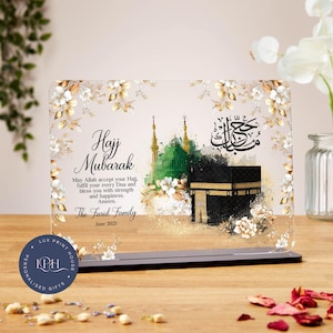 May include: A clear acrylic plaque with a black base, featuring the words "Hajj Mubarak" and a message. The design includes images of a mosque and the Kaaba, with floral accents. The text "The Farid Family June 2025" is also present.