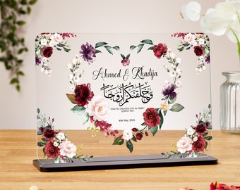 May include: A clear acrylic wedding sign with a black base. The sign features the names "Ahmed & Khadija" and Arabic calligraphy, surrounded by a floral heart design with burgundy, white, and purple flowers. The date "16th May 2024" is also included.