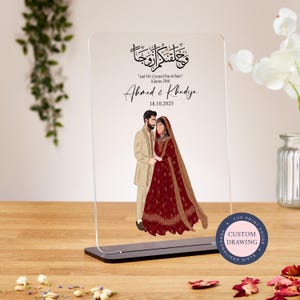 Puede incluir: Un soporte de acrílico transparente con una ilustración personalizada de una pareja con atuendo tradicional de boda. El soporte presenta el texto árabe "و حلفتكم الزوجا" y la cita "Y os creamos en parejas" (Corán 78:8). Los nombres "Ahmed & Khadija" y la fecha "18.10.2025" también están incluidos. El soporte está sobre una base negra y tiene una pegatina circular con el texto "LUX PRINT HOUSE PERSONALISED GIFTS CUSTOM DRAWING".