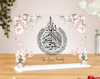 Può includere: Una placca in acrilico trasparente con un motivo floreale e un'iscrizione in calligrafia araba nera. La placca ha una base bianca e il testo "The Ejaz Family" è stampato sotto la calligrafia.
