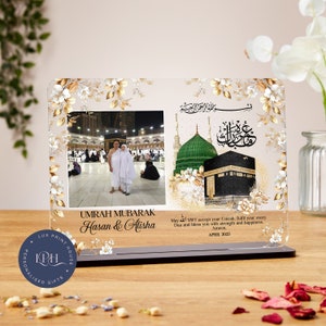 Pode incluir: Uma colagem de fotos emoldurada em branco e dourado com duas fotos. A foto superior mostra a Caaba em Meca, Arábia Saudita. A foto inferior mostra duas pessoas com túnicas brancas em pé em frente à Caaba. O texto na moldura diz "Umrah Mubarak Hasan & Alisha" e "Que Allah SWT aceite sua Umrah, cumpra cada uma de suas Duas e abençoe você com força e felicidade. Amém. Abril de 2025."