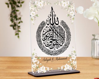 Può includere: Una placca in acrilico trasparente con un motivo floreale nero e un'iscrizione in calligrafia araba nera. L'iscrizione è circondata da un bordo floreale bianco. La placca ha un supporto nero e il testo "Aaliyah & Muhammad" è stampato in basso.