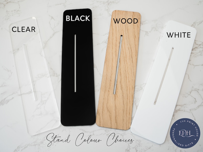 May include: Four acrylic stands in different colors: clear, black, wood, and white. Each stand has a slot for holding a phone or tablet. The stands are displayed on a white marble surface. The text "Stand Colour Choices" is written below the stands.