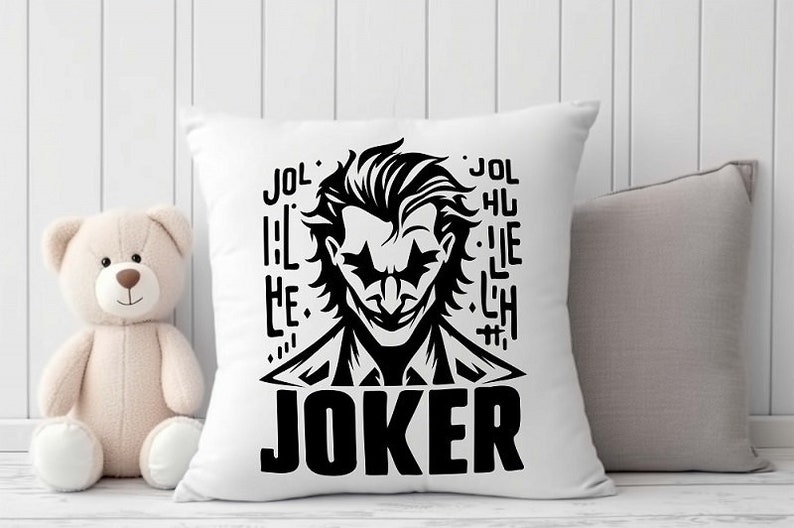 Puede incluir: Almohada blanca con una ilustraci&oacute;n en blanco y negro del Joker de Batman. La ilustraci&oacute;n incluye la palabra "JOKER" en letras grandes y el texto "JOL HU LIE LH" en letras m&aacute;s peque&ntilde;as.