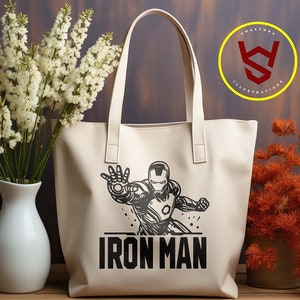 May include: Cream-colored tote bag featuring a black and white Iron Man graphic. The bag has two handles and the words "IRON MAN" printed below the character. The image also includes a logo and file type options.