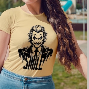 Puede incluir: Una camiseta amarilla con una ilustraci&oacute;n en blanco y negro de un personaje de Joker sonriente. El texto "SMILE" est&aacute; impreso debajo del personaje.