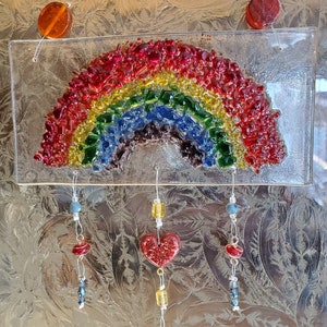 Op de afbeelding: Een glazen regenboog-zonenvanger met een rood, oranje, geel, groen, blauw en paars ontwerp. De regenboog is gemaakt van kleine stukjes glas. De zonenvanger hangt aan een touwtje met drie bedels: een rood hart, een rode kraal en een gouden tag met het woord "Happy" erop.