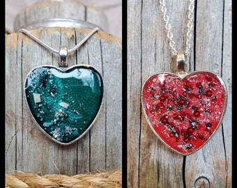 May include: Two silver heart-shaped pendants on chains. The pendant on the left is green with white flecks, and the pendant on the right is red with black and white flecks.