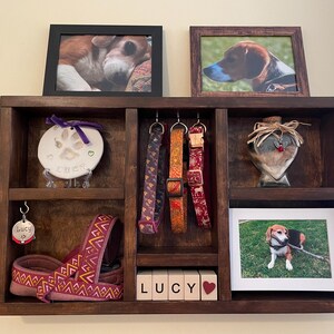 May include: A dark brown wooden pet display shelf with framed photos, dog collars, a paw print keepsake, and a dog harness. The shelf includes the name "LUCY" in wooden blocks and a heart.